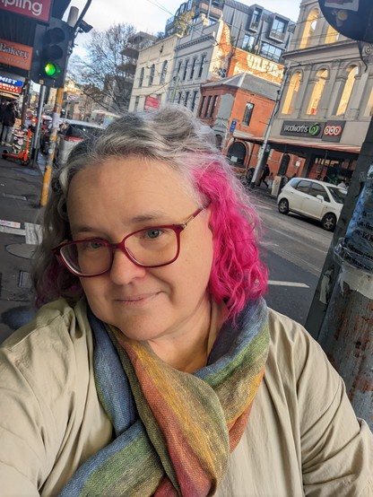 Selfie of a person with curly silver & pink hair, a rainbow linen scarf, & pink glasses