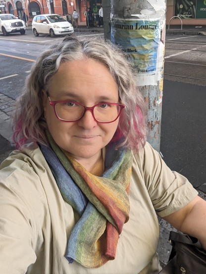 Selfie of a person with curly silver & pink hair, a rainbow linen scarf, & pink glasses