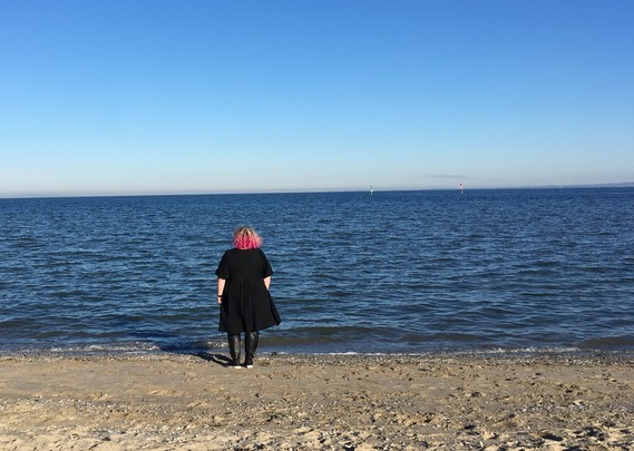 Rear view of a person in a black dress standing on a beach looking at the horizon