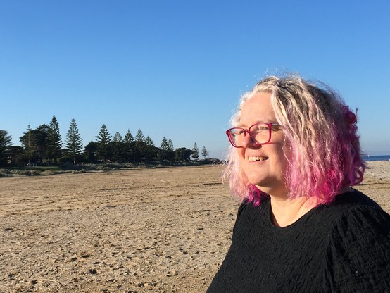 Smiling person standing on a beach. In the background are the same trees visible in the behind-the-scenes shot from the FNF video
