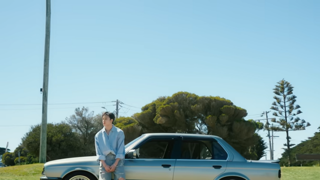 Bang Chan leaning on a vintage car parked on a grassy area