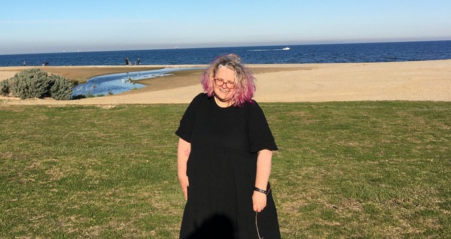 Person in a black dress & pink hair, leaning on an imaginary car while holding a STICK