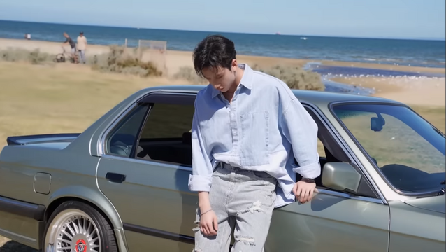 Bang Chan leaning on a vintage car, looking down at his feet. Behind him is a beach with scrubby grass