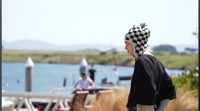 Hyunjin in black & white checkered beanie with earflaps. A marina is blurry visible behind him