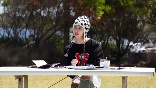 Hyunjin sitting at a picnic table with painting materials in front of him