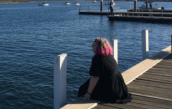 A person in a black dress with pink hair, sitting on the edge of the boardwalk