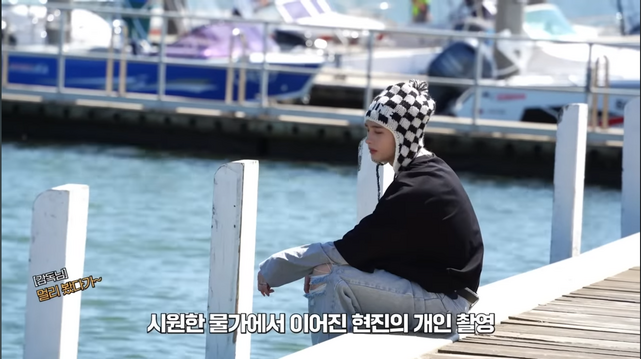 Hyunjin sitting on the edge of the boardwalk with his legs over the edge. The marina is visible behind him.