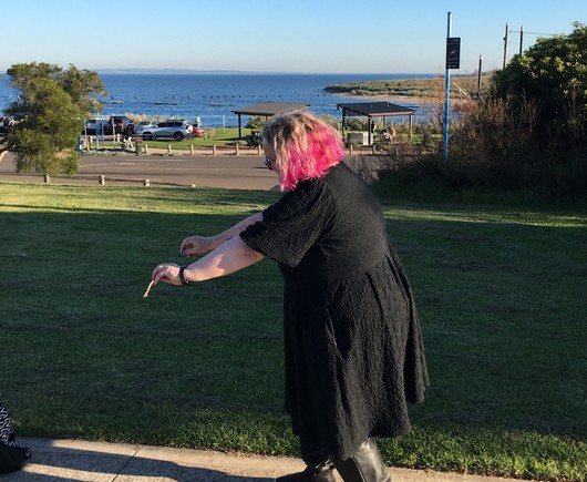 A person with pink hair & wearing a black dress, riding their imaginary bike along a waterfront path