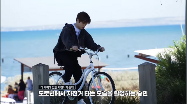 Seungmin riding a bike along a waterfront path, with some picnic shelters behind him