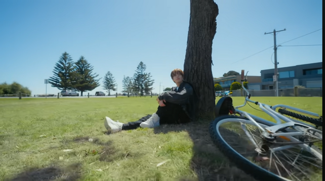 Seungmin sitting under a tree with his back against it. A bike is on its side next to him