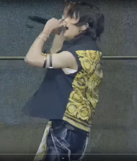 Hyunjin on stage wearing a mostly black outfit with a yellow-ish scarf attached to his right hip