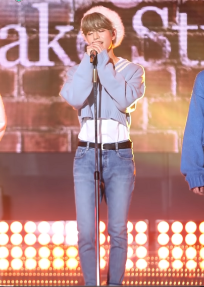 Seungmin standing on a stage performing, while wearing a soft blue cropped sweater and a fuzzy beret. His eyes are partly closed and he's wearing sparkly pink eyeshadow 