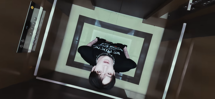 Bang Chan stands in an elevator and tilts his head back to look into the camera that is directly above him