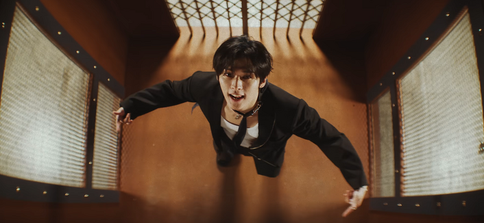 Lee Know sings and spreads out his arms close to the sides of an old elevator while he looks up at the camera filming him from directly above